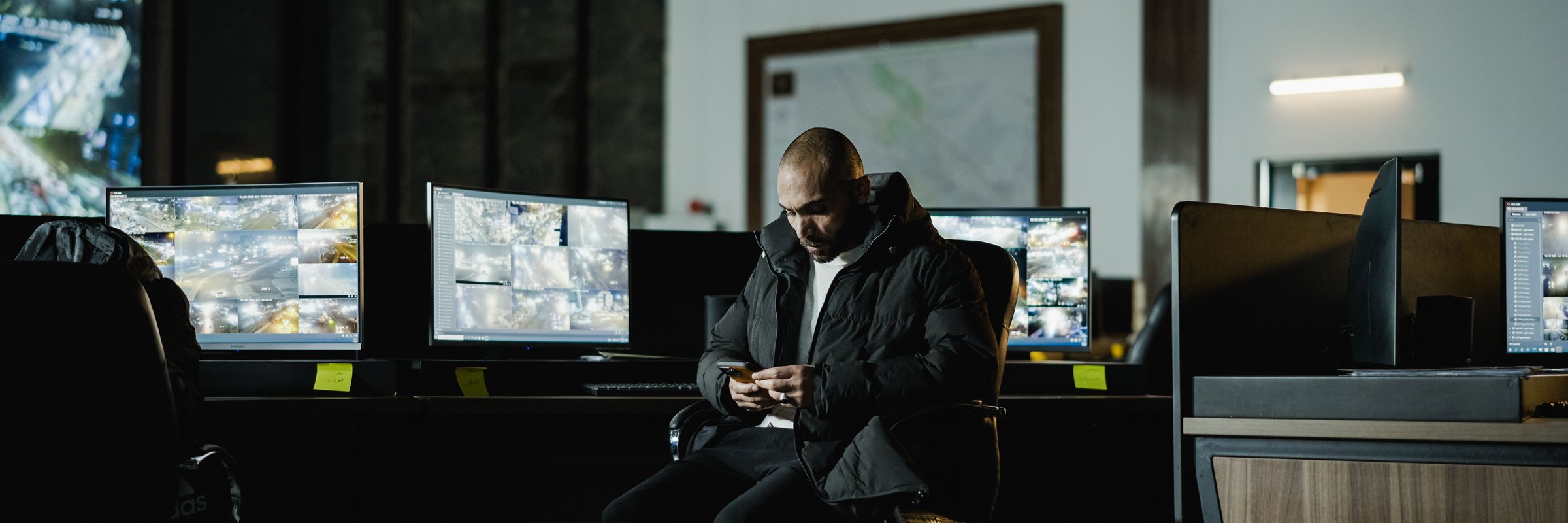 Oficial de segurança sentado em sala de controle escura, observando várias telas de vigilância.